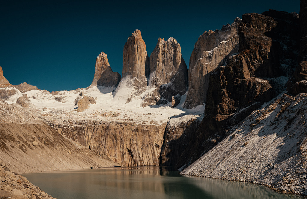 The three magestic golden towers of Torres del Paine against a deep blue sky with Lago Torres in the foreground.