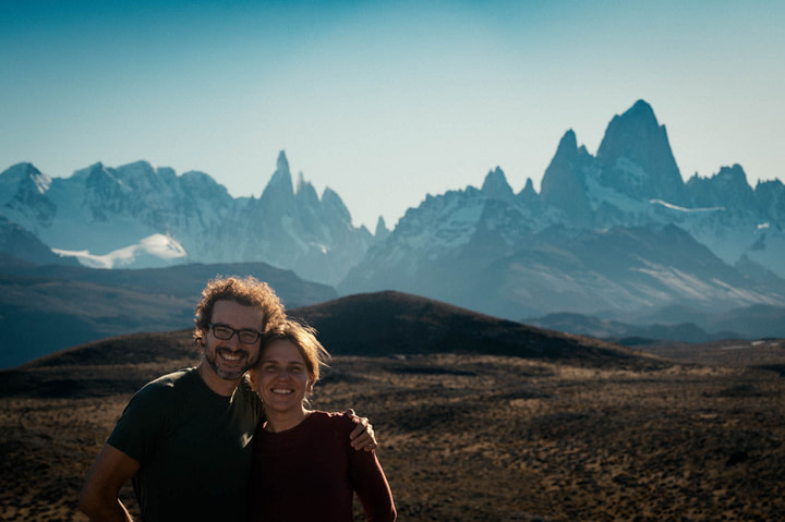 Theo and Maxine standing in front of the Fitz Roy Mountain range. Mountains are vast and the show various shades of blue.